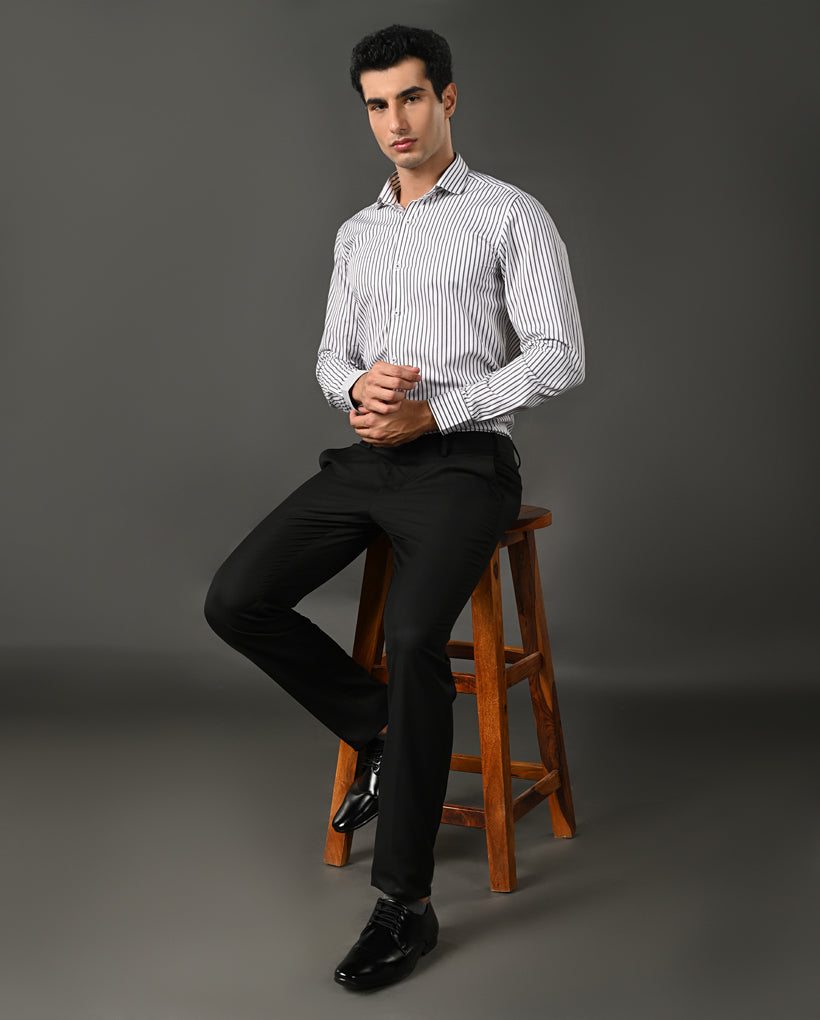 Man sitting on a wooden stool wearing a striped shirt and black pants against a gray background