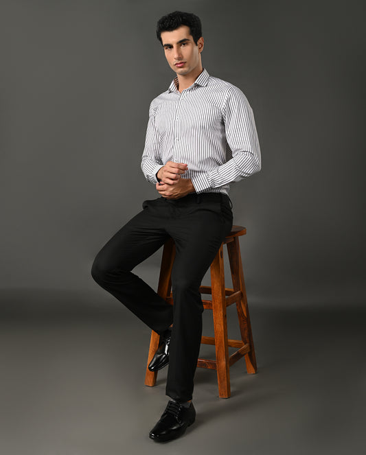 Man sitting on a wooden stool wearing a striped shirt and black pants against a gray background